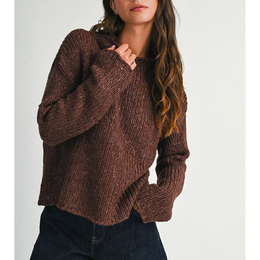 A woman wearing a chunky brown knit sweater and dark denim shorts, standing against a plain light gray background.